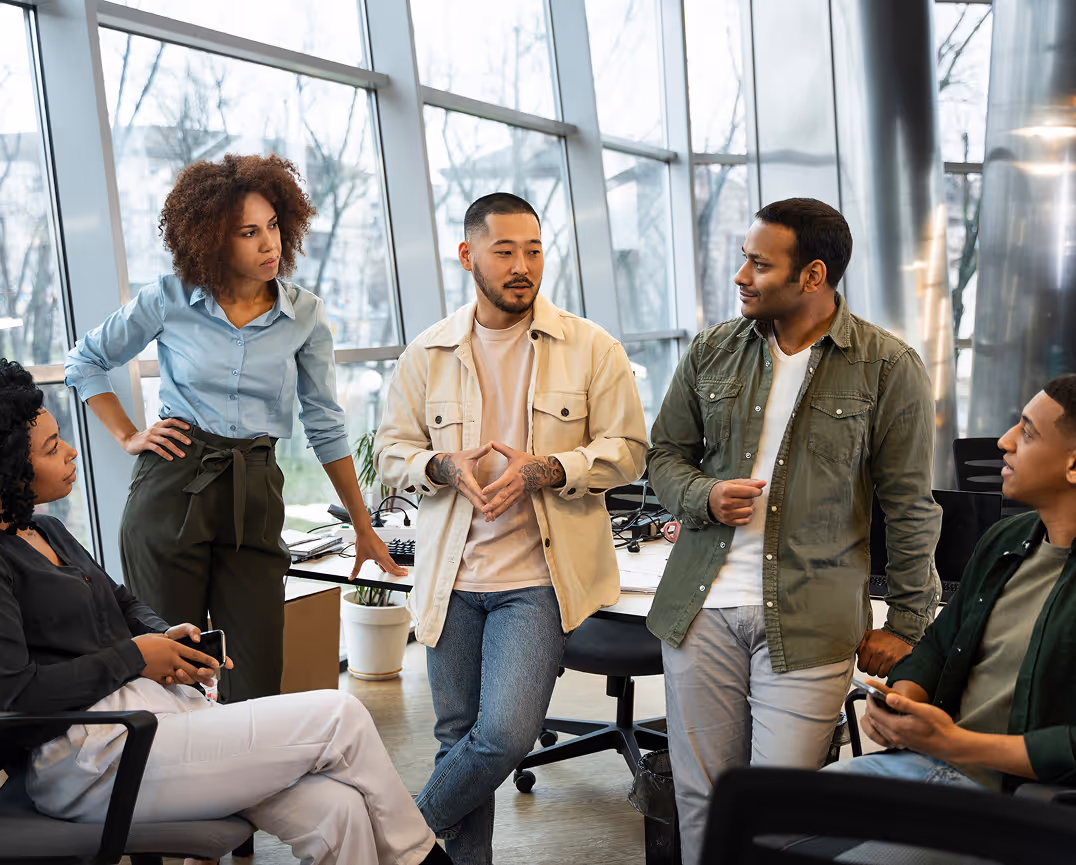 Five diverse coworkers engaged in a discussion inside a modern office with large windows.