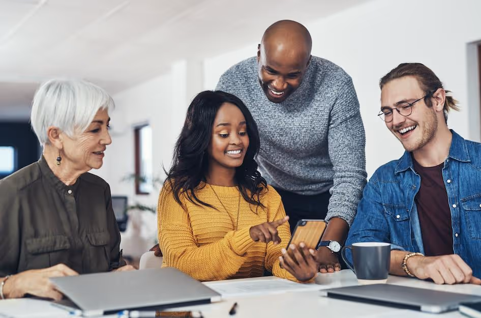 Diverse group of four young professionals smiling and looking at a smartphone together at a bright office table.