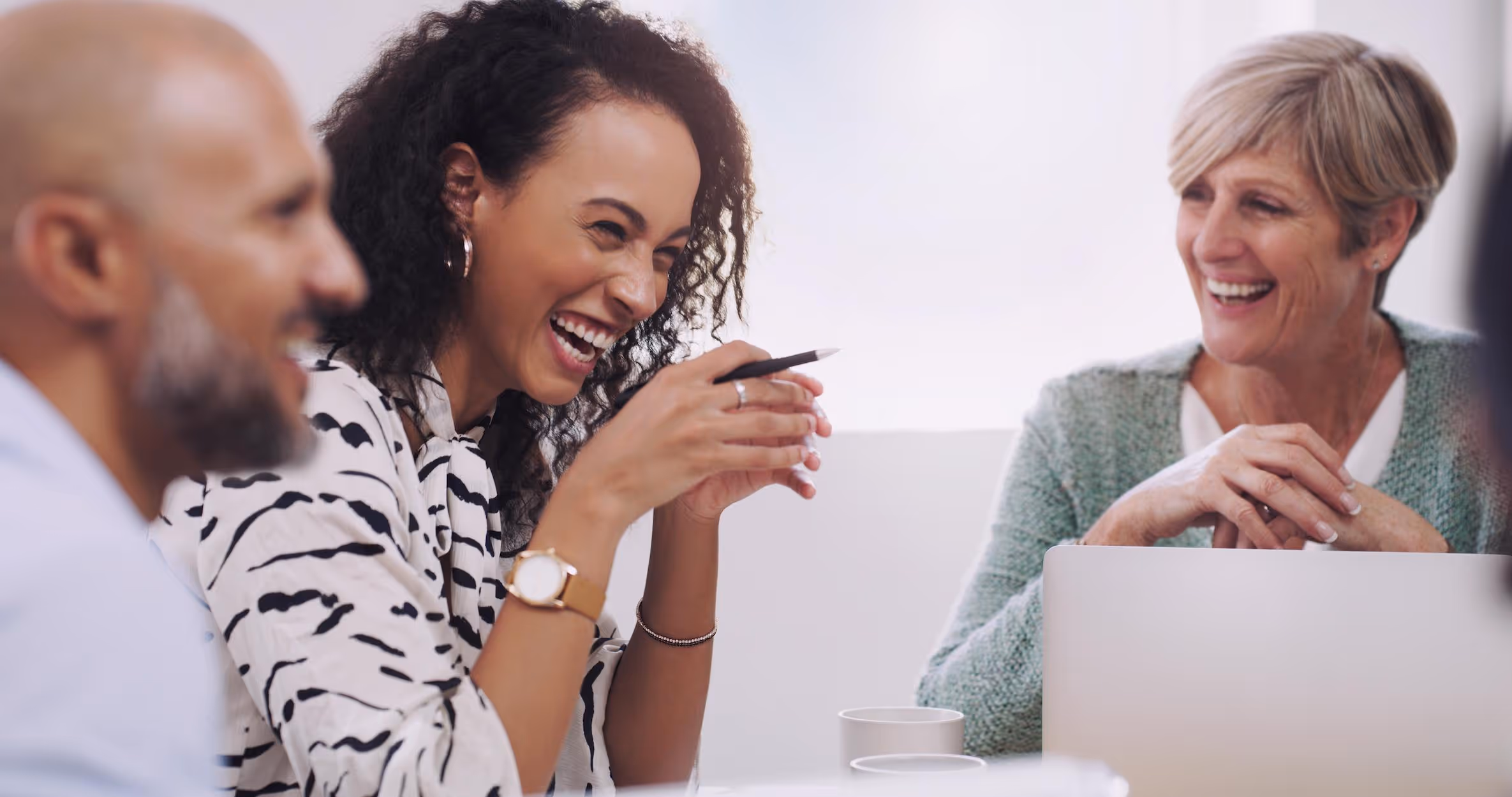 Three coworkers laughing and enjoying a meeting around a table in a bright office.