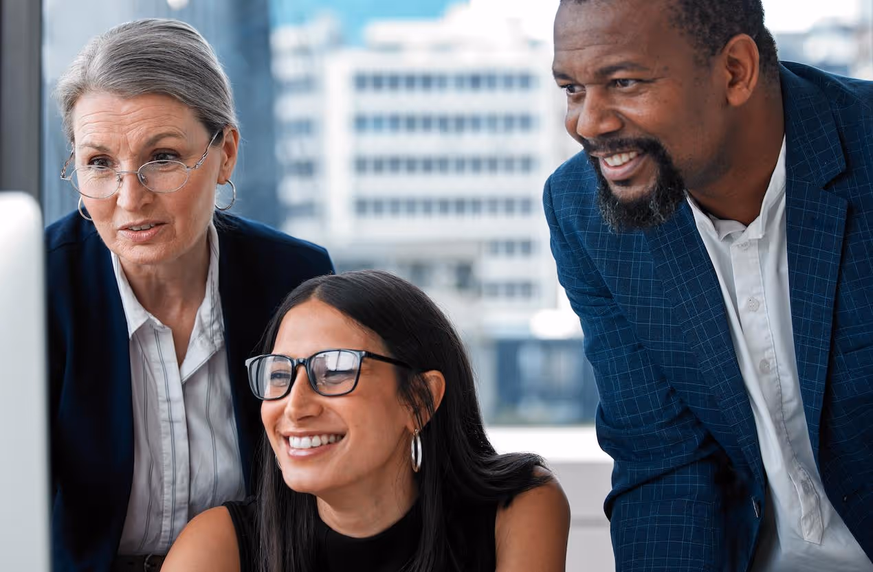 Three diverse business professionals smiling and discussing work in a modern office with cityscape background.