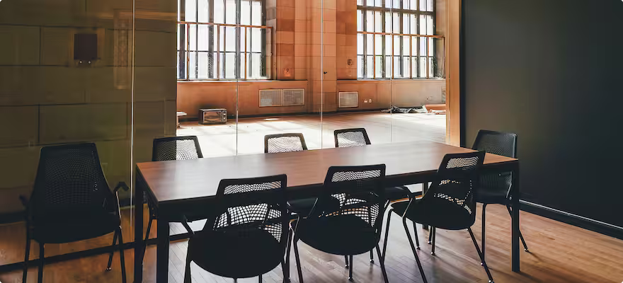 Empty conference room with a wooden table surrounded by black mesh chairs and large windows reflecting in a glass partition.