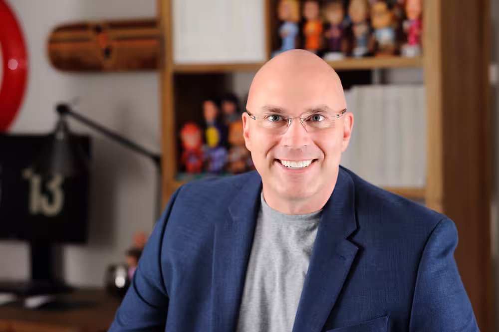 Smiling bald man wearing glasses, a gray shirt, and a blue blazer in front of a bookshelf with figures and books.