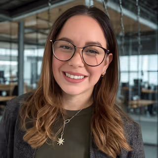 Smiling woman with long brown hair, glasses, and star-shaped necklace in a modern office setting.