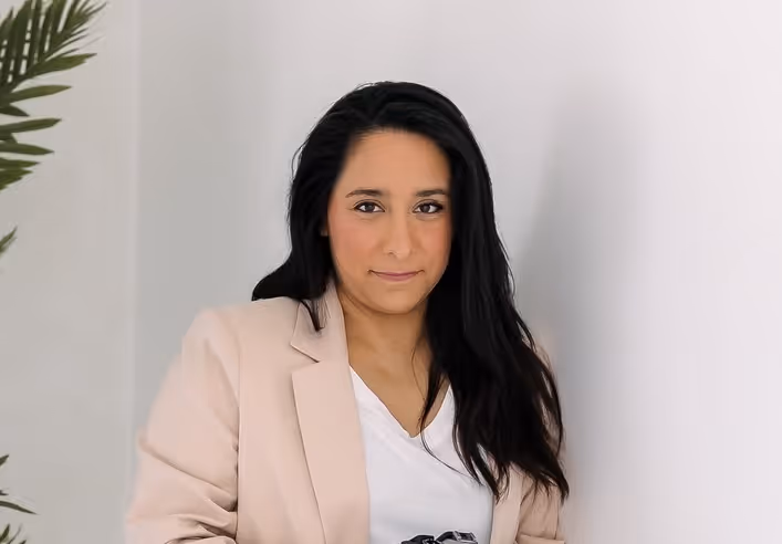 Woman with long dark hair wearing a beige blazer and white top, standing indoors near a white wall and a green plant.