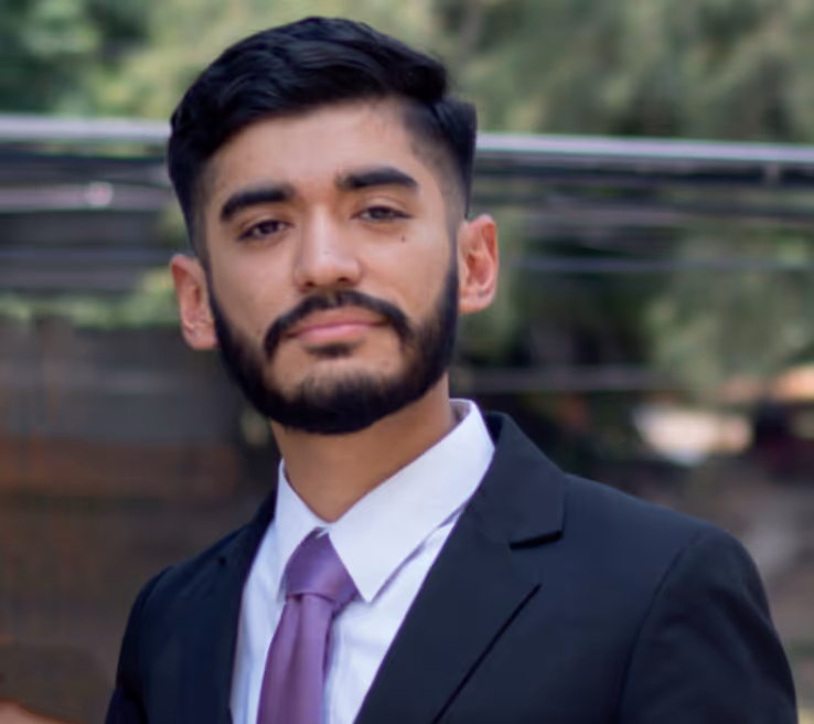 Portrait of a young man with dark hair and beard wearing a black suit and purple tie outdoors.