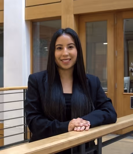 Woman with long black hair smiling, wearing a black blazer, standing indoors with hands resting on a wooden railing.