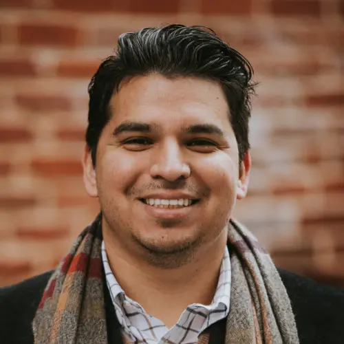 Smiling man with dark hair wearing a plaid shirt, scarf, and dark jacket against a blurred brick wall.