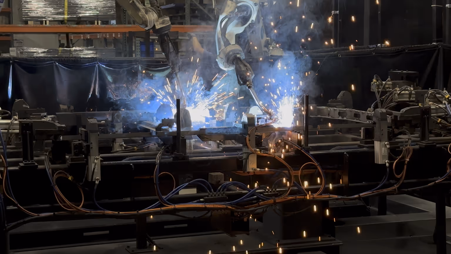 Automated robotic arms welding metal parts with sparks and blue light in an industrial fabrication workshop.