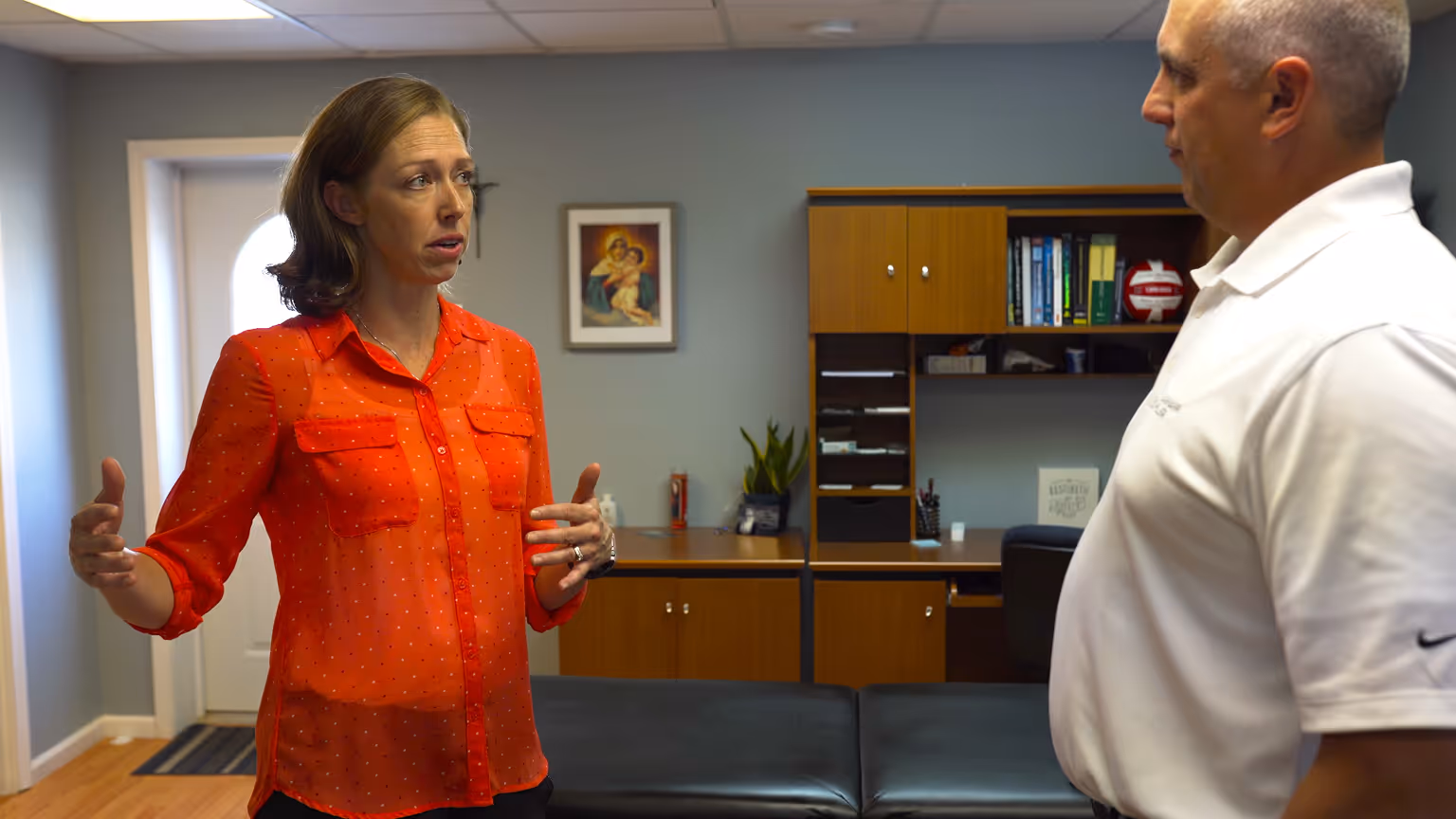 Two adults having a conversation in a well-lit office with wooden cabinets and a framed picture on the wall.