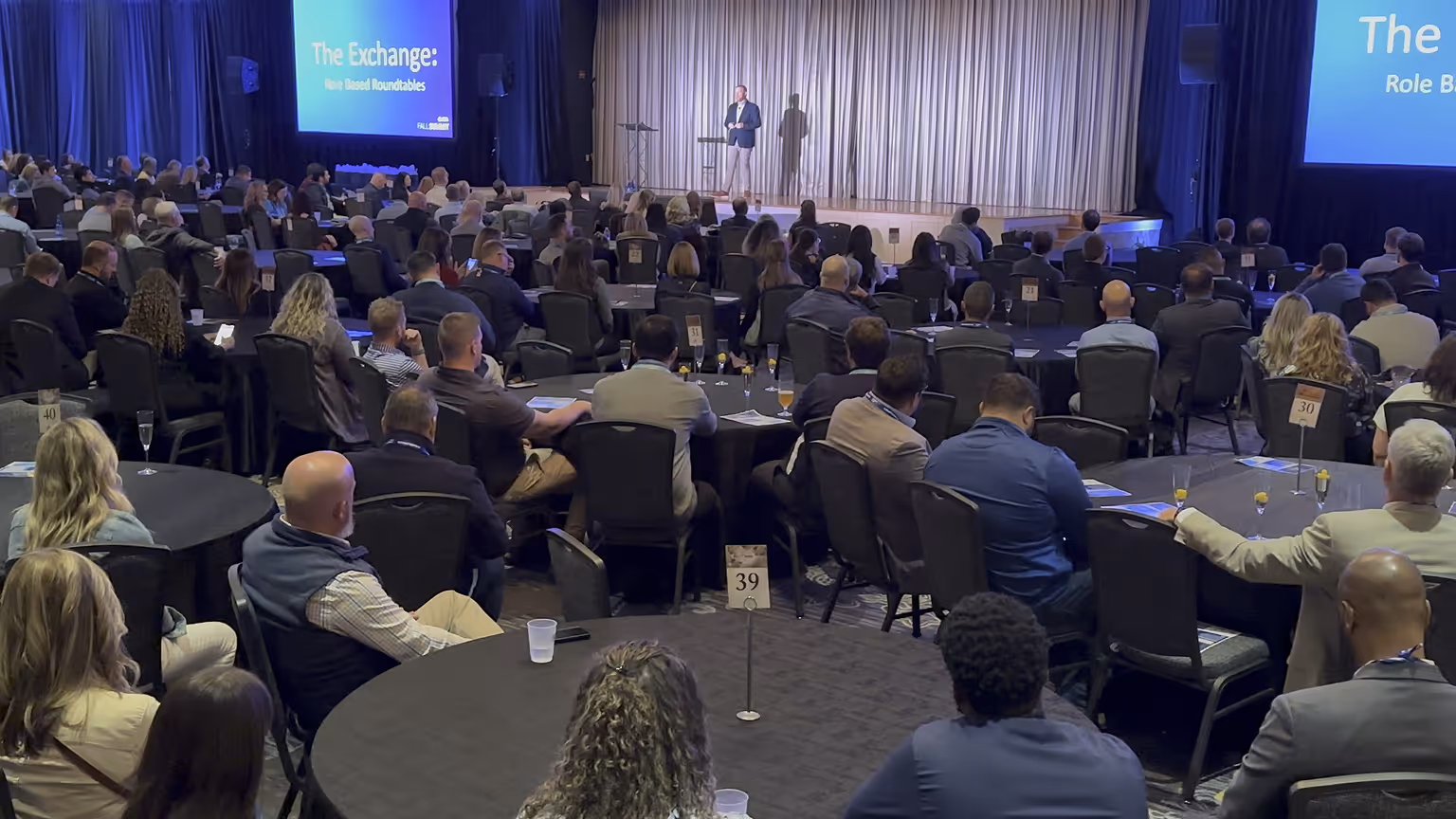Audience seated at round tables listening to a speaker on stage during a conference event titled 'The Exchange: Role Based Roundtables'.