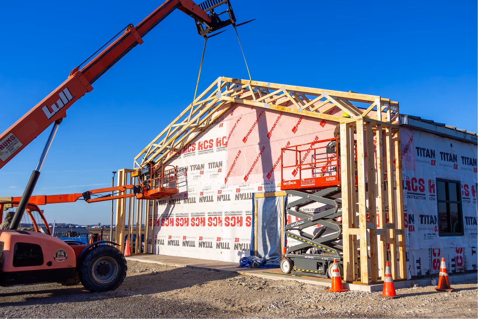 Construction site with a partially framed building wrapped in protective sheeting, a crane lifting materials, and an orange scissor lift.