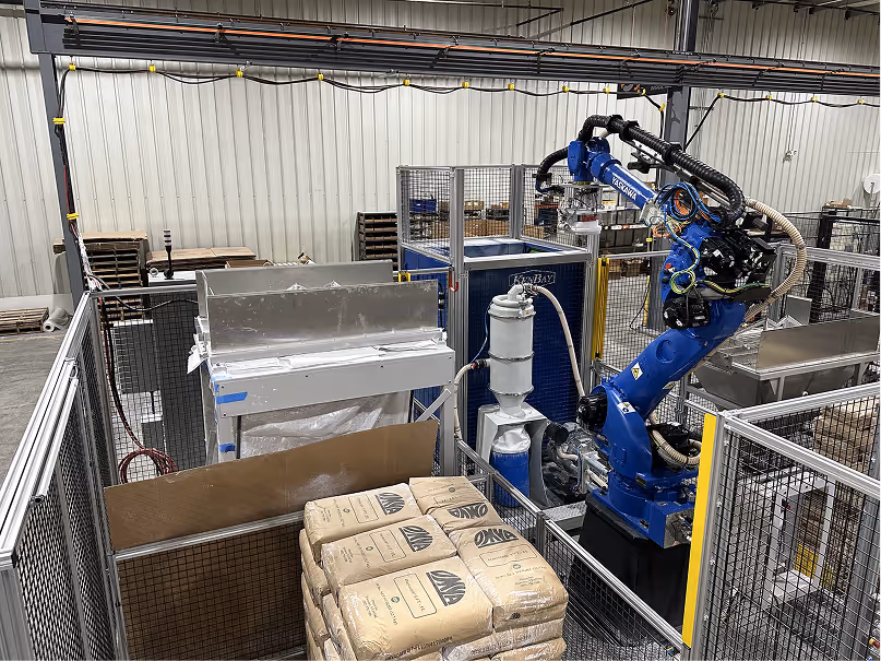 Industrial setting with a blue Yaskawa robotic arm inside a fenced area, surrounded by equipment and stacked bags of materials.