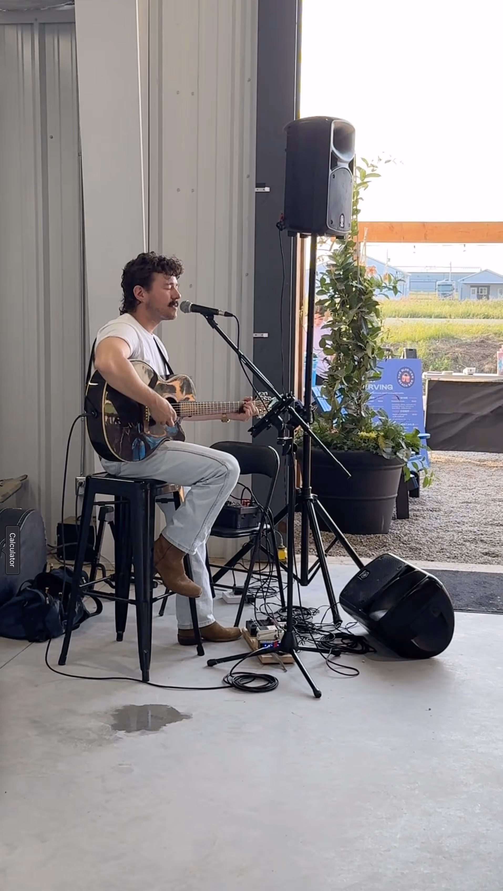 Person sitting on a stool singing and playing the guitar. A mic stand and speaker are infront.
