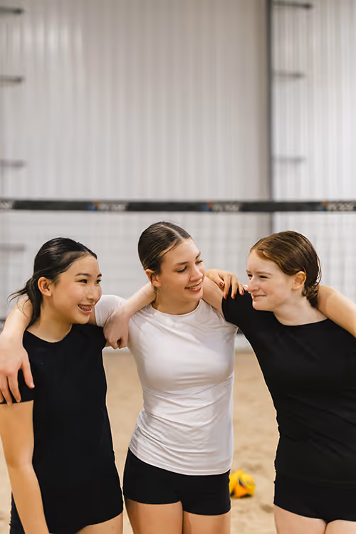 Three young female volleyball players in athletic wear standing with arms around each other in an indoor sand court, smiling.