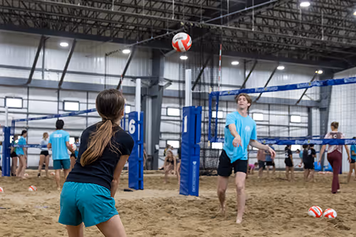 Young people playing indoor beach volleyball on sandy courts with nets and blue posts.
