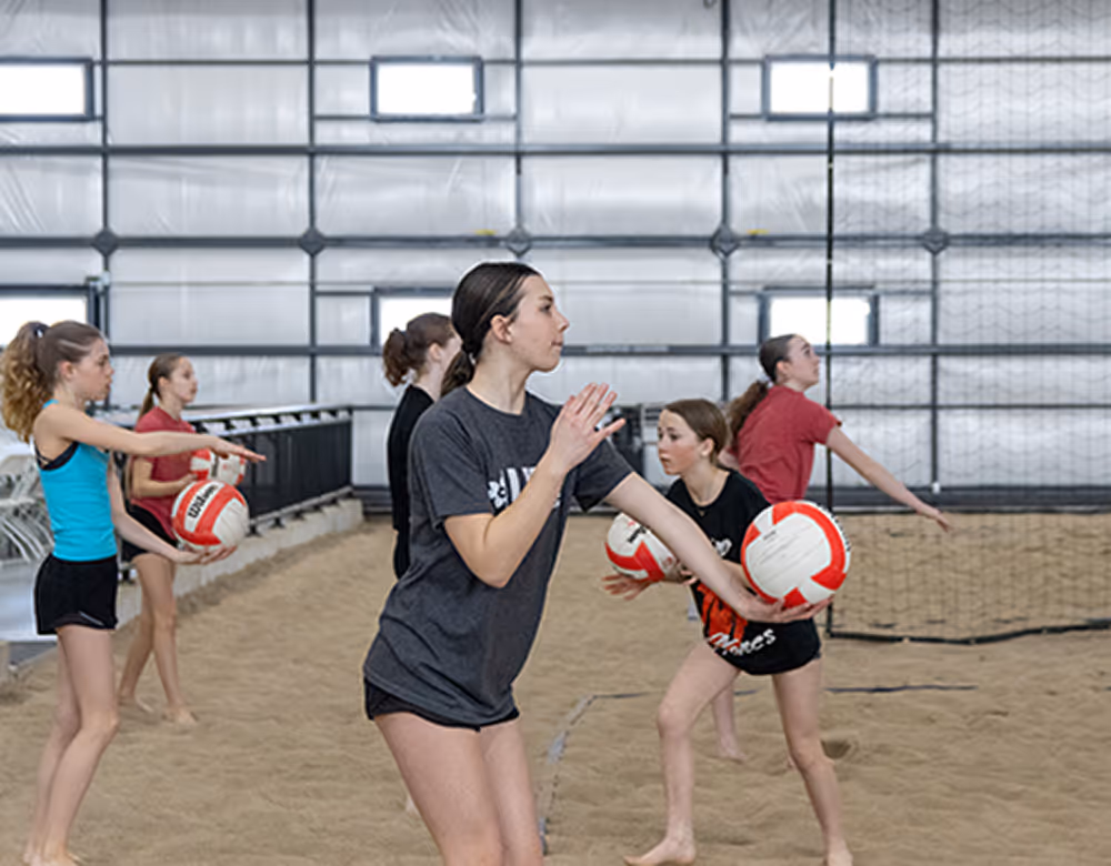 Group of young athletes practicing volleyball indoors on a sand court.