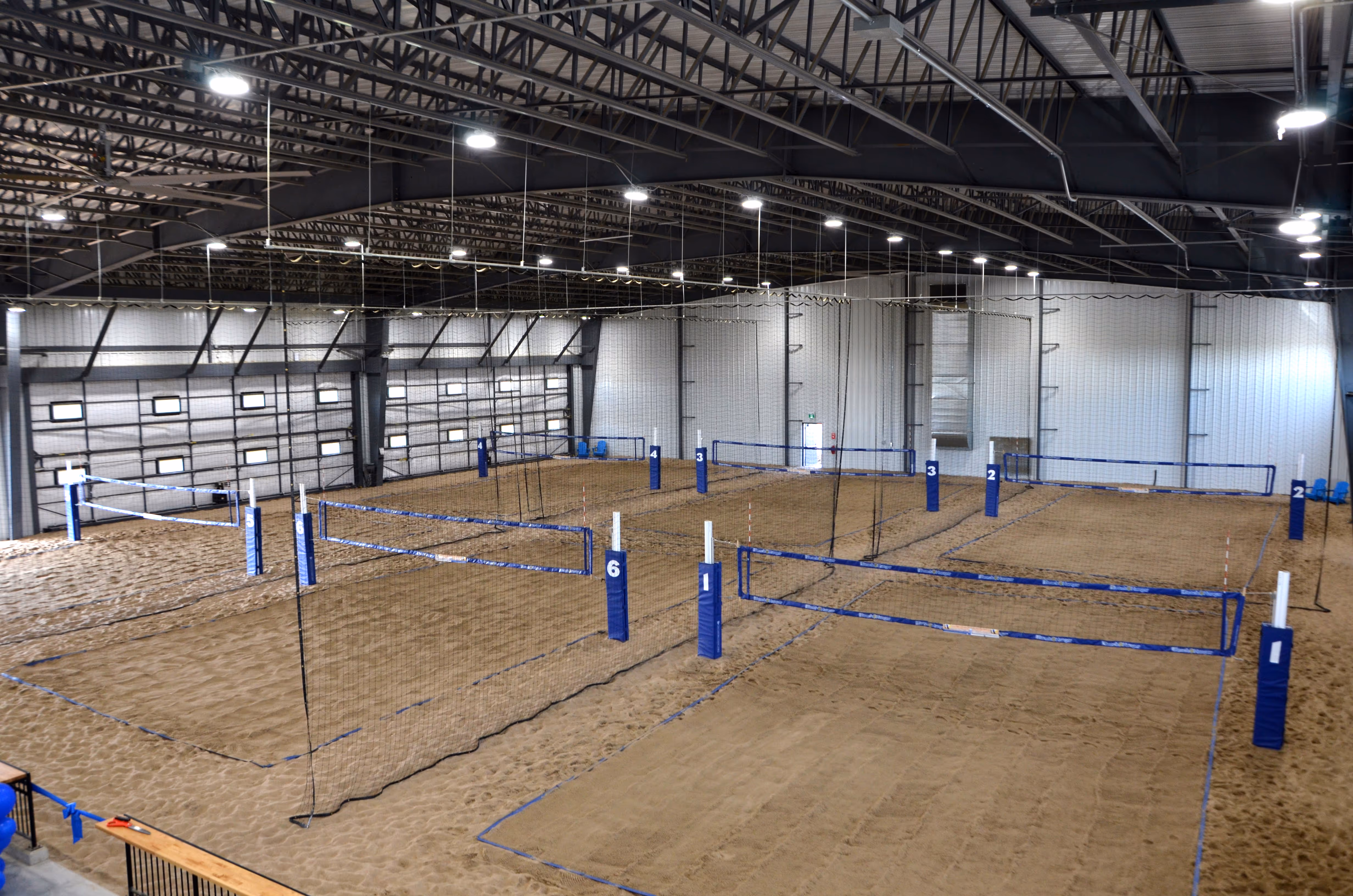 The Beach Hangar indoor beach volleyball courts with sand, blue and white net posts, and overhead lights in a large warehouse-style building.