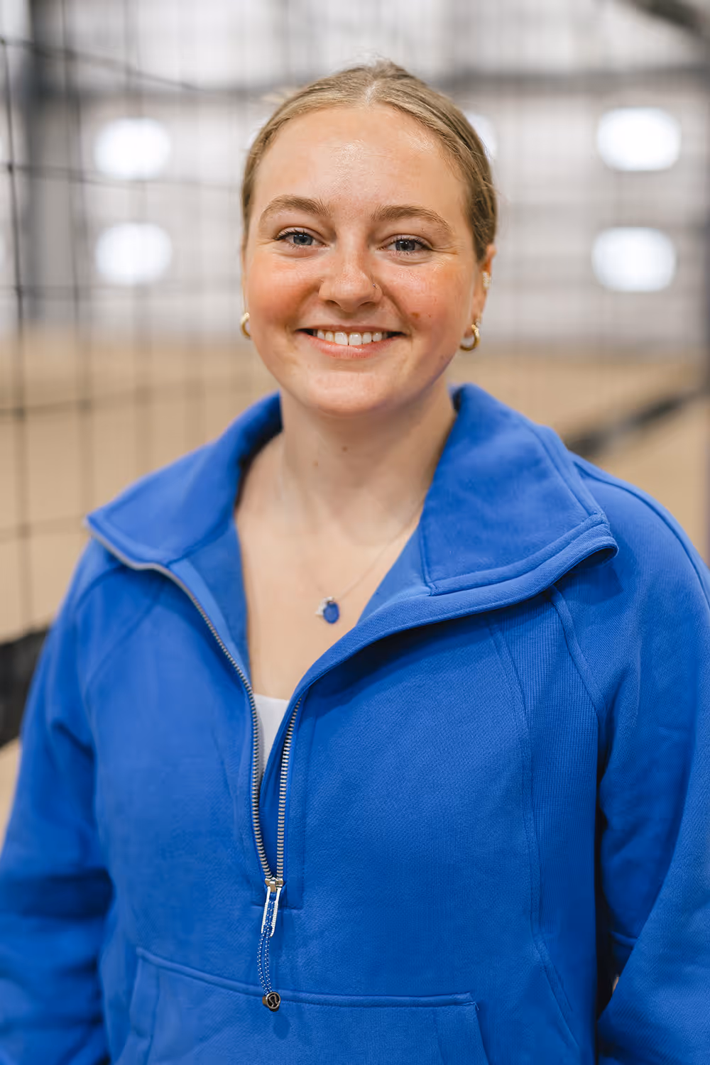 Erin, Beach Hangar's head coach, smiling wearing a blue zip-up jacket and a blue pendant necklace in an indoor sports setting.