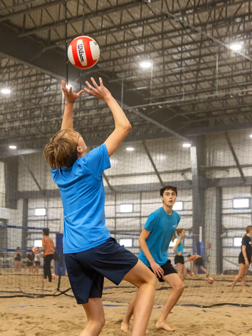 Two young men playing indoor beach volleyball with a red and white Wilson volleyball, one preparing to set the ball.