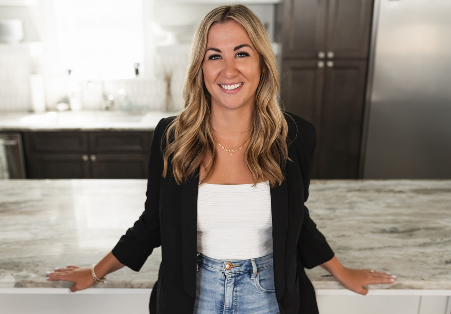 Smiling woman with blonde wavy hair wearing a black blazer and white top, leaning on a marble kitchen island.
