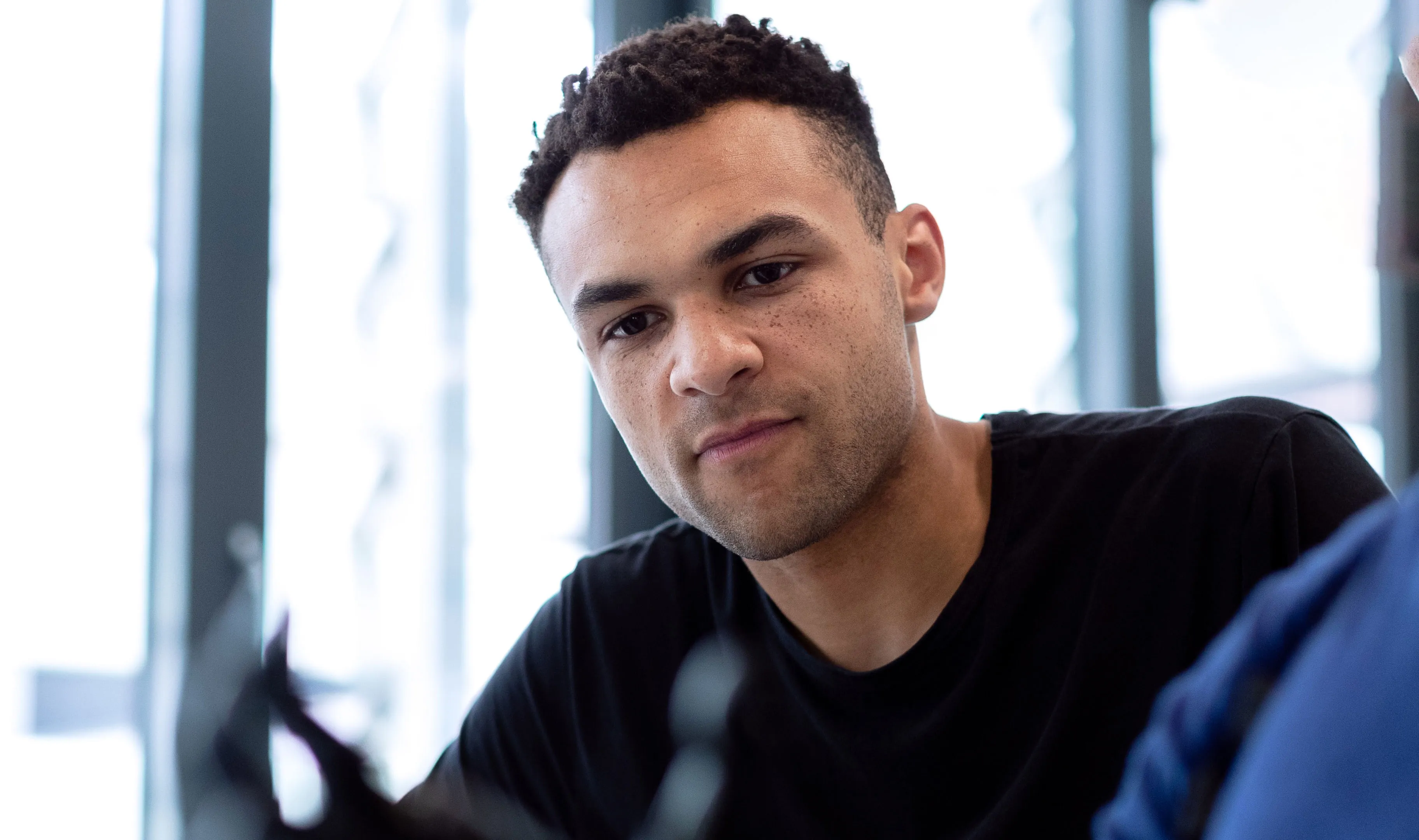 Young man with short curly hair and freckles looking intently at something, wearing a black shirt, indoors with large windows in the background.