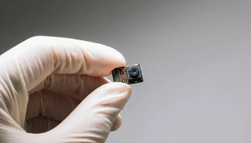 Hand wearing a white glove holding a small electronic camera module with a visible lens and circuitry.