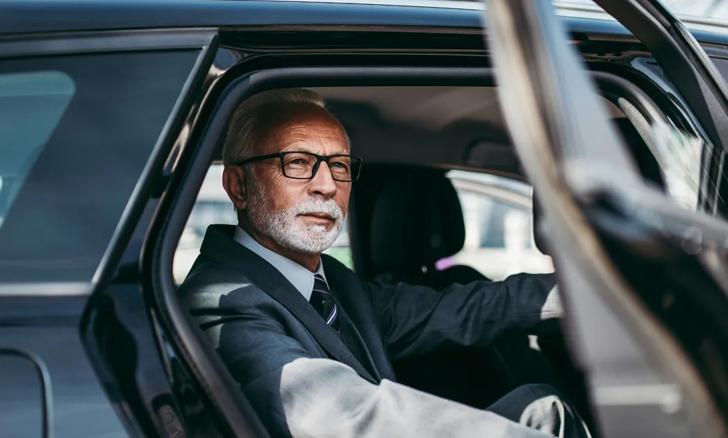 Older man with gray hair and beard wearing glasses and a suit sitting in the driver's seat of a black car.