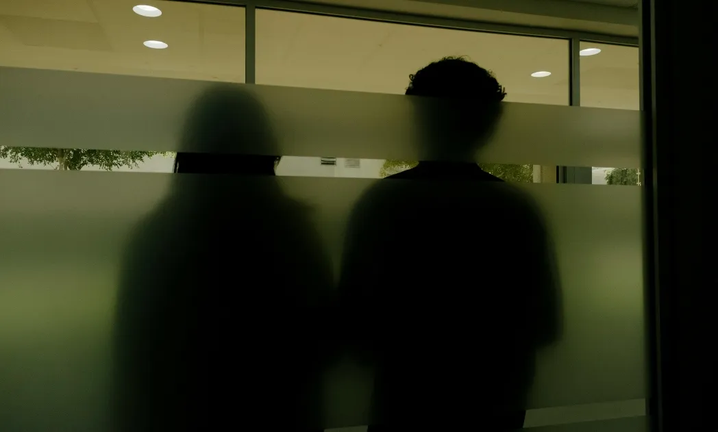 Two blurred silhouettes of people standing behind frosted glass with horizontal clear stripes.