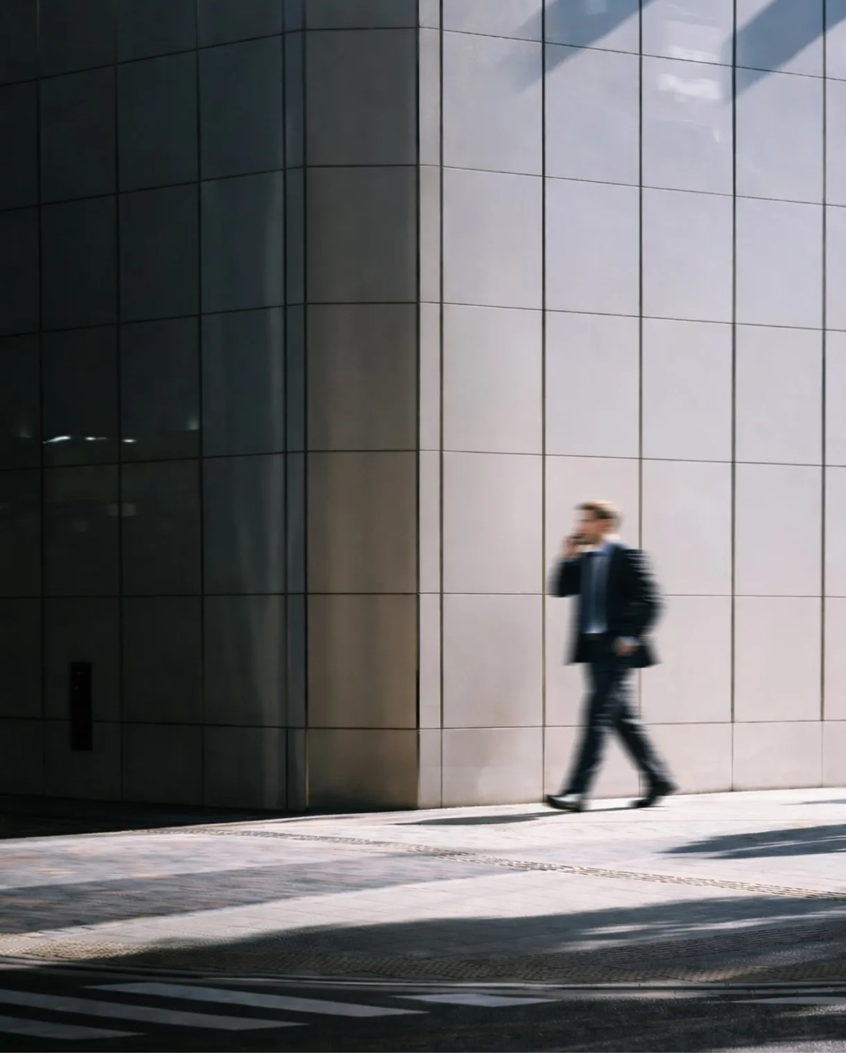 Blurred silhouette of a businessperson walking and talking on a phone beside a tiled urban building.