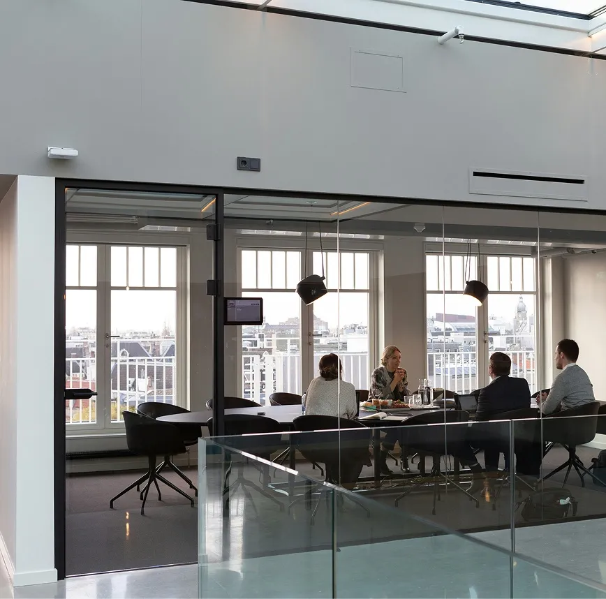 Four people seated around a conference table in a glass-walled office with large windows overlooking a cityscape.