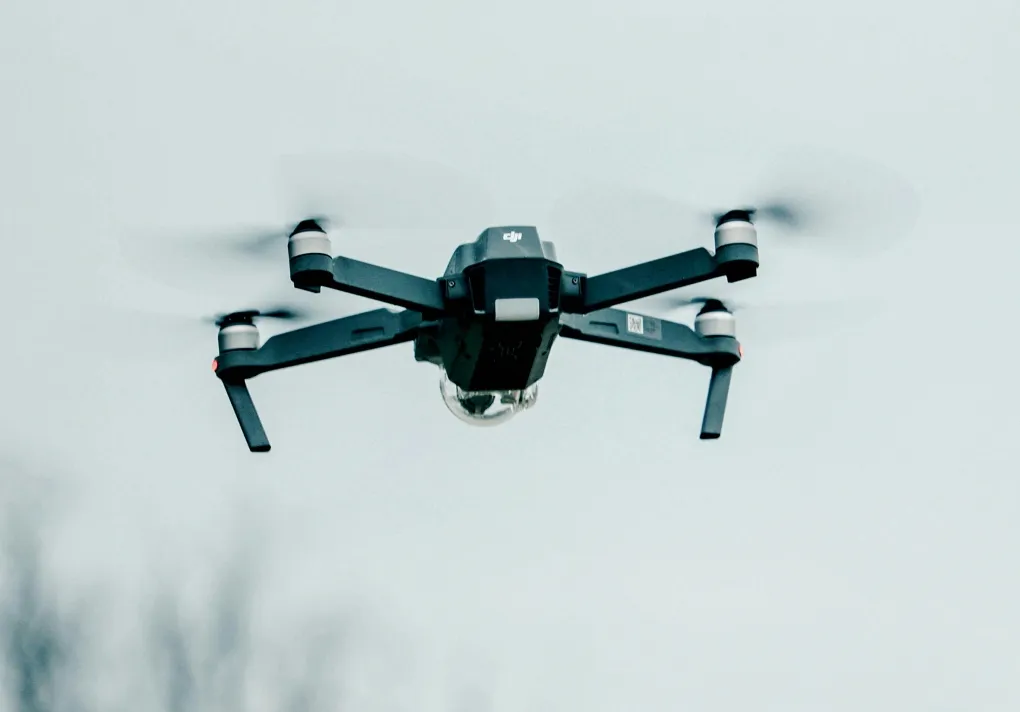 Black quadcopter drone flying against a pale sky with blurred spinning propellers.