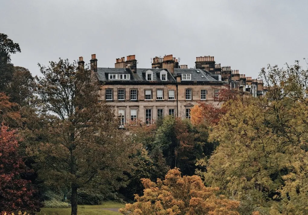 Large stone building partially obscured by trees with autumn foliage under a gray sky.
