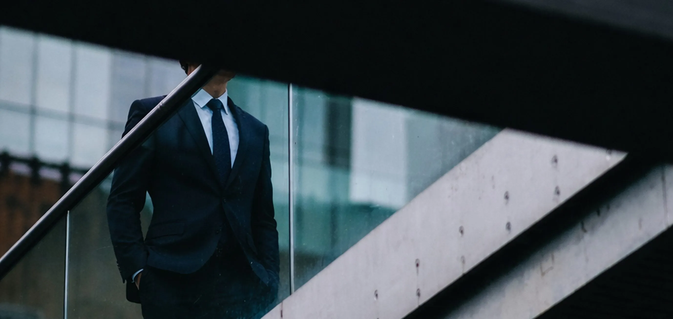 Man in a dark suit with a tie standing behind glass panels and concrete beams in an urban setting.