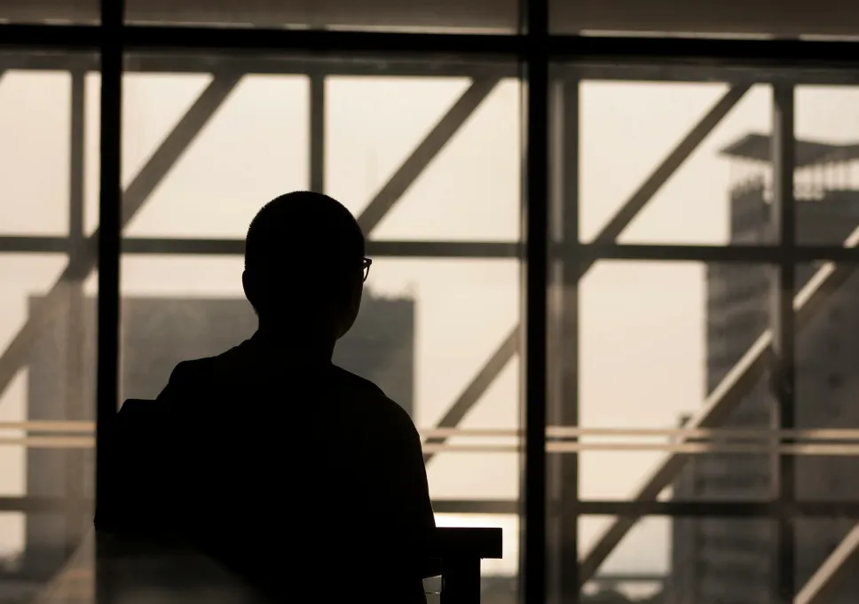 Silhouette of a person wearing glasses sitting indoors facing large windows with an urban skyline in the background.