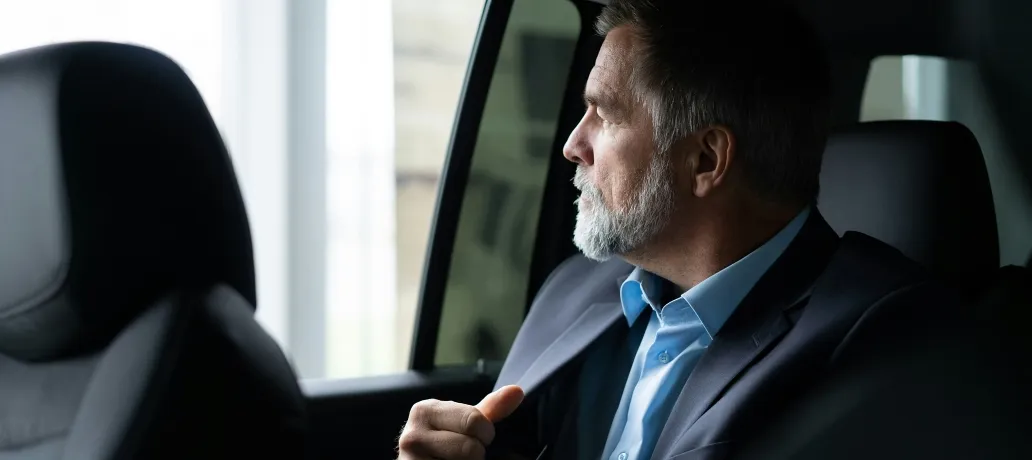 Older man with gray hair and beard wearing glasses and a suit sitting in the driver's seat of a black car.