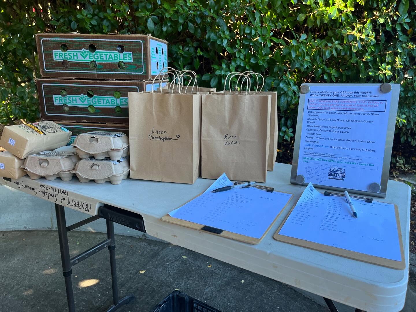 A Shakefork pickup site set up: produce boxes, egg cartons, named paper bags, two sign-in clipboards and the printed final-week share list carrying the early-renewal offer