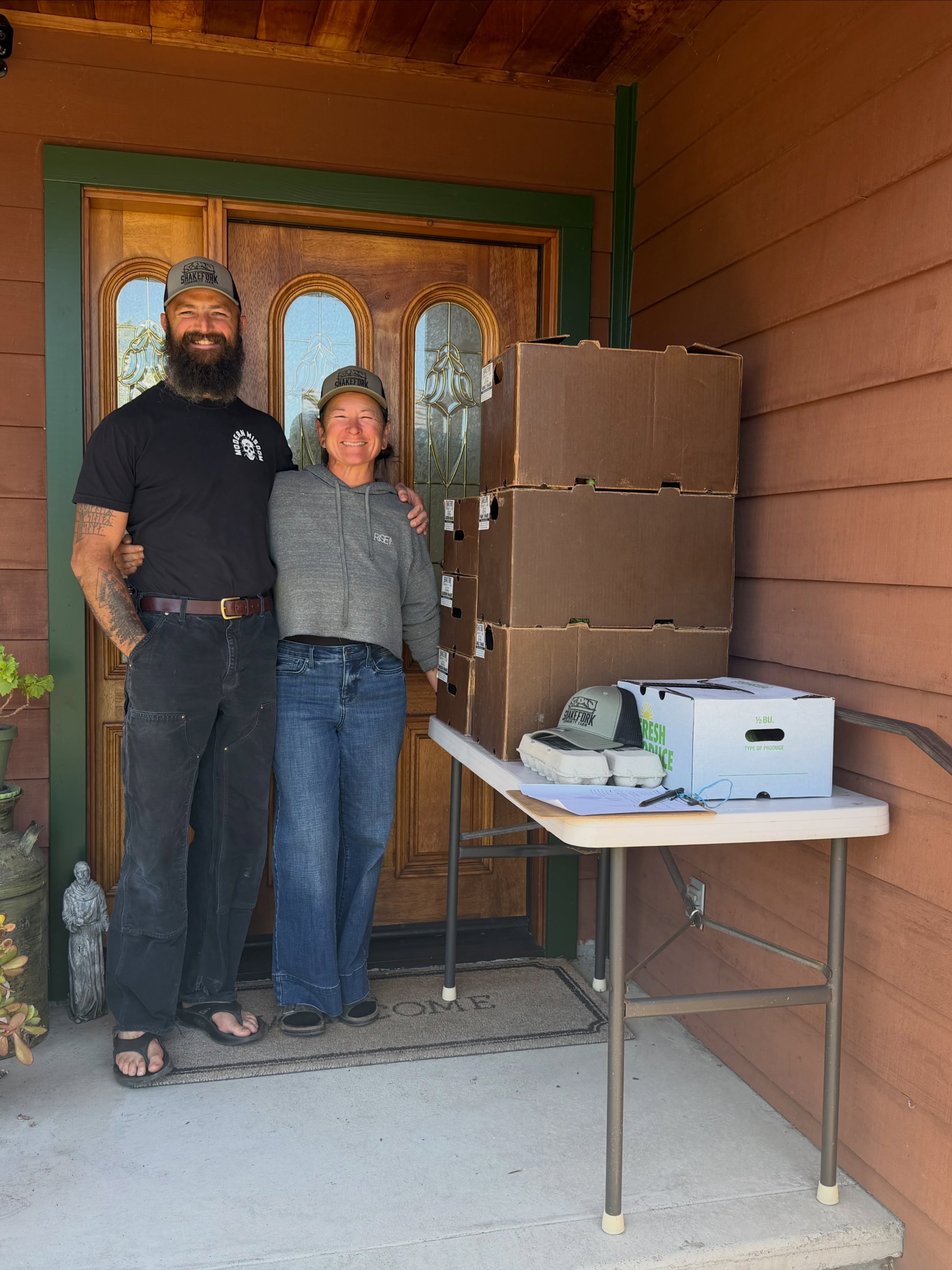 Melanie and Kevin together at a porch pickup site, produce boxes and egg cartons stacked on the table beside them