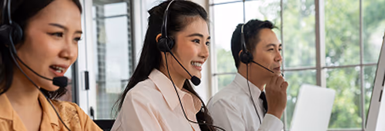 Three customer service representatives wearing headsets, working at desks in a bright office with large windows