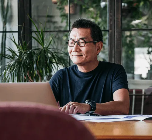 Smiling man wearing glasses and a black t-shirt, sitting at a desk with papers and a laptop, working in a bright room with large windows and green plants in the background.