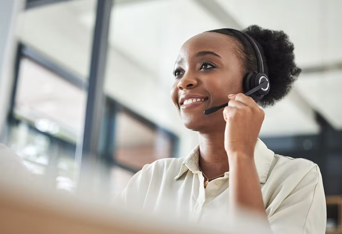 A young Black woman with a pleasant expression works in a call center. She is wearing a headset with a microphone and smiling while looking up and to her right.