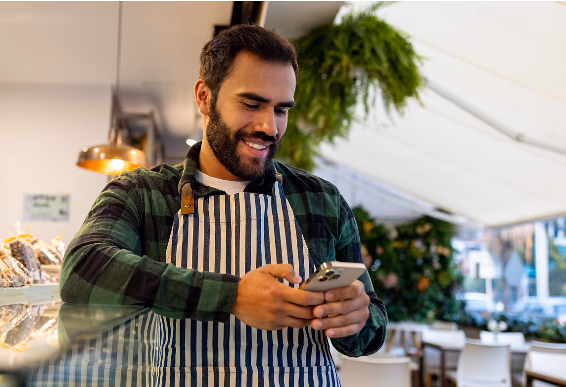 Smiling man wearing a striped apron in a café, holding a smartphone and looking at the screen.