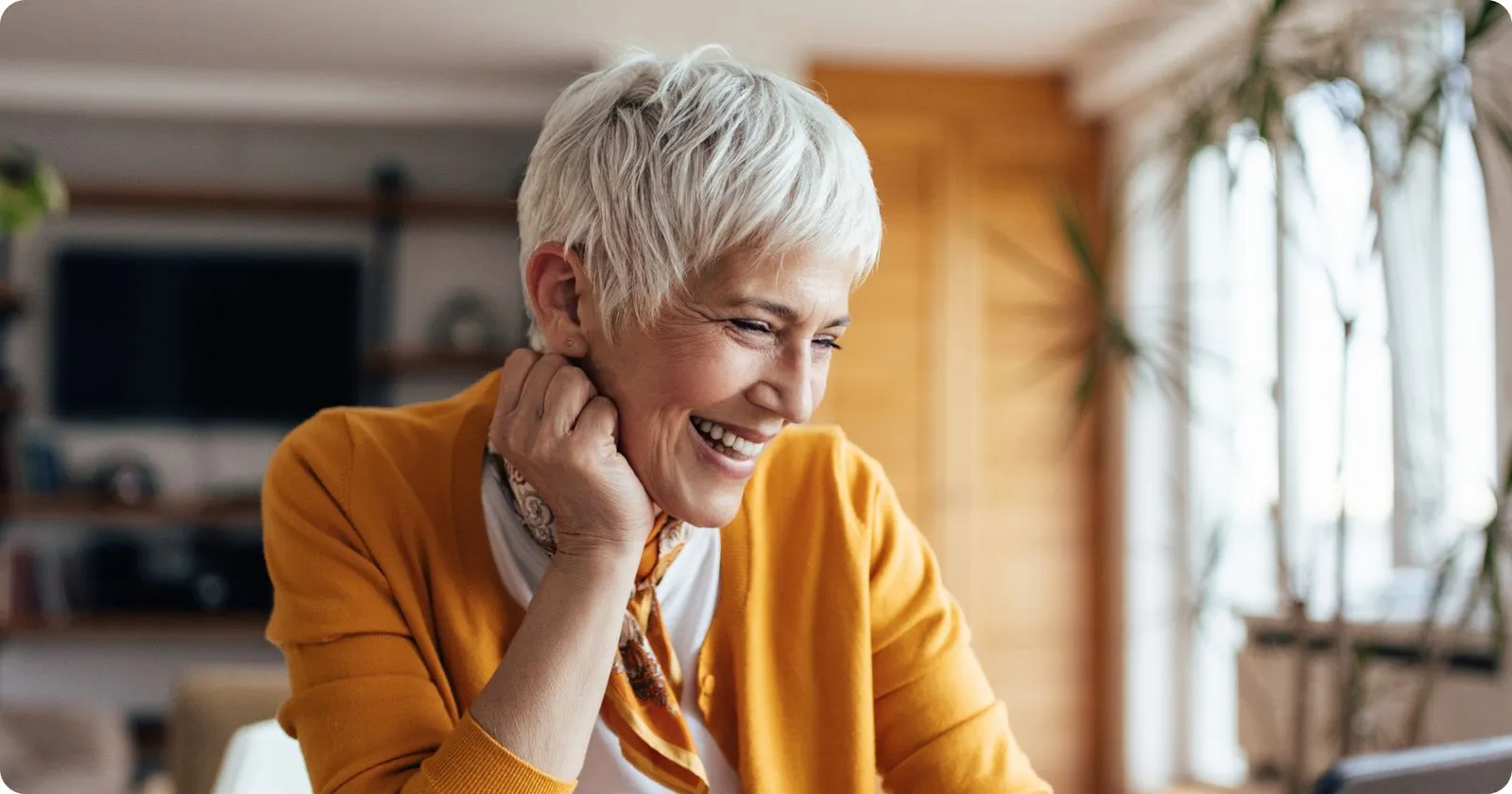 Smiling older woman in an orange cardigan looking at a computer in a bright living room.