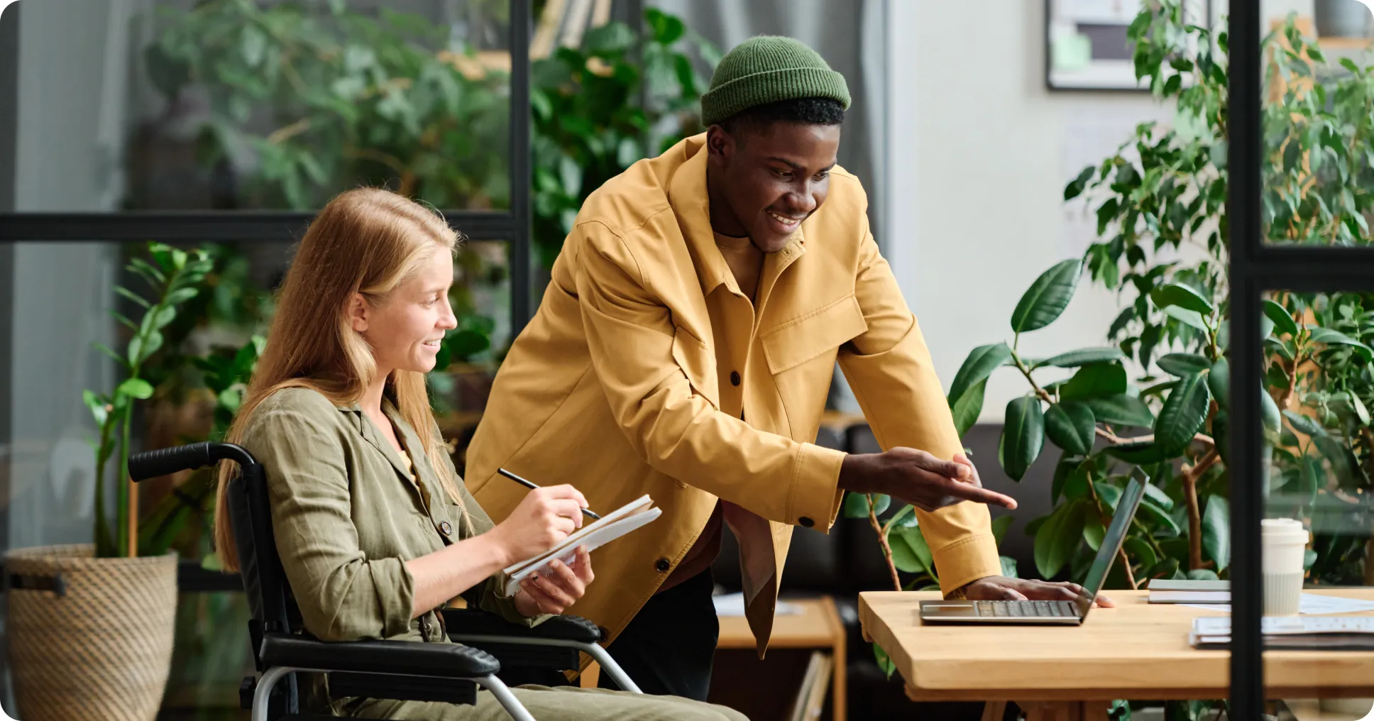 Man and woman collaborating at a desk with a laptop in a bright office filled with plants.