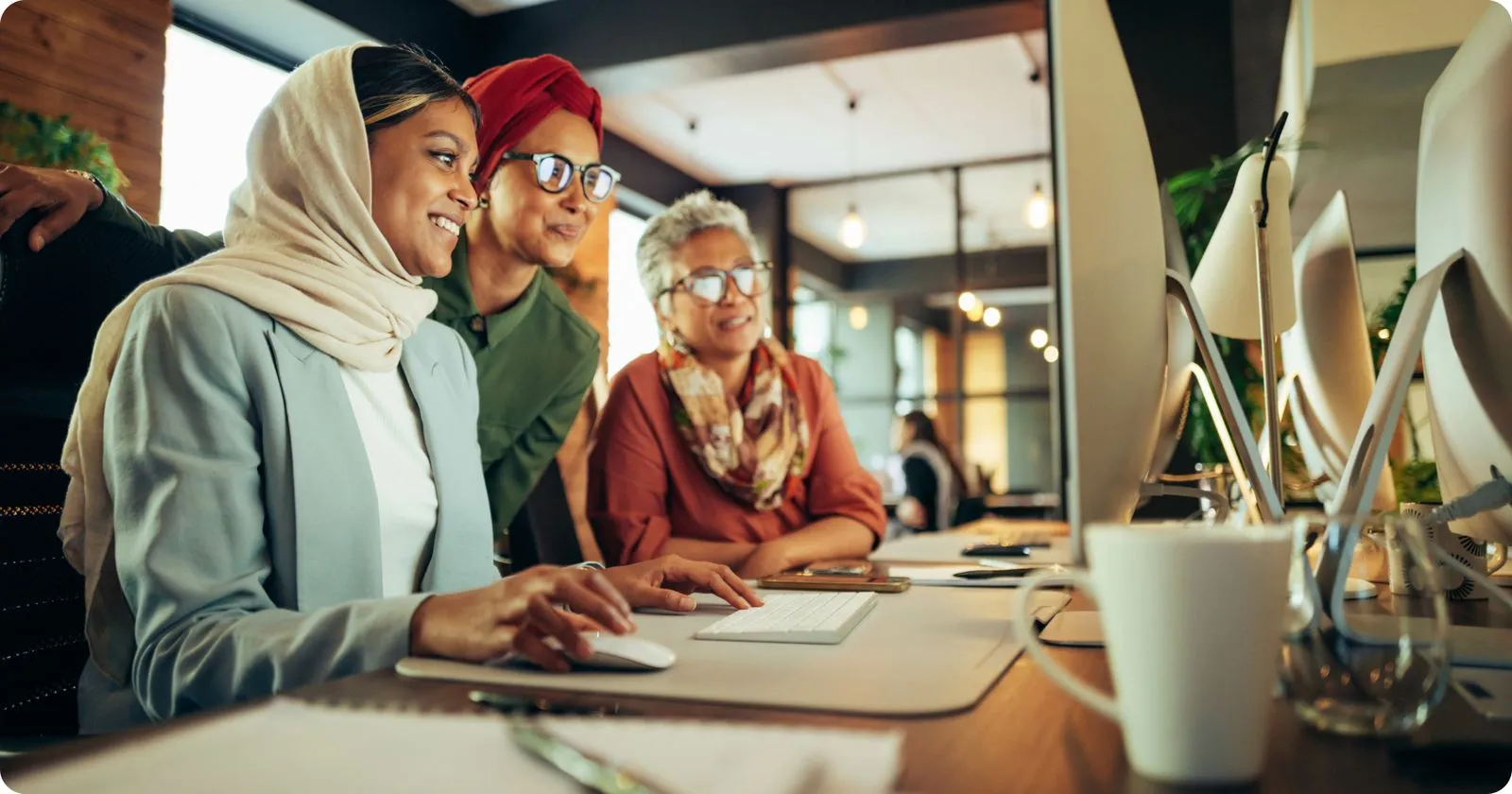 Three women smiling and collaborating at a computer in a modern office workspace.