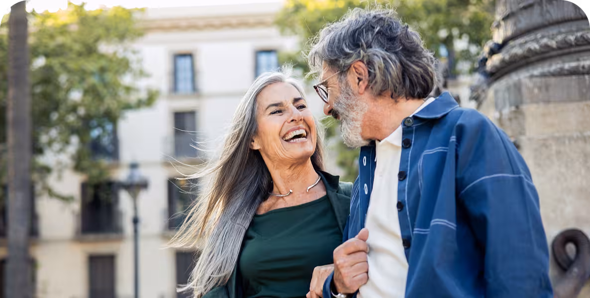 A smiling older woman with long, grey hair looks at and laughs with an older man with grey hair and a beard, both outdoors.