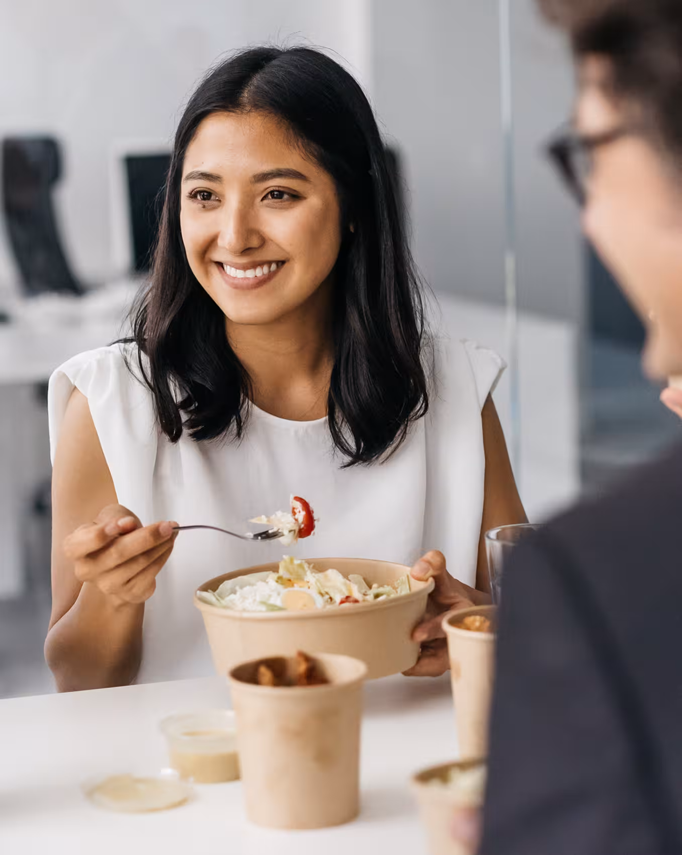 A close-up of a smiling woman with dark hair in a white top eating a salad from a bowl, looking towards someone off-camera in an office environment.