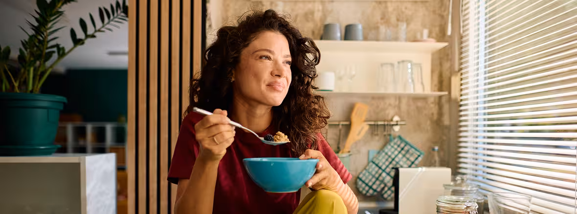 A woman with long, curly dark hair sits at a kitchen counter, eating from a blue bowl with a spoon and looking thoughtfully out a window with blinds.