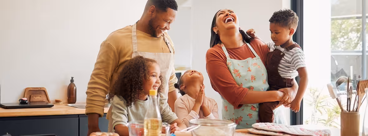 A joyful family of five in a kitchen, with parents and three children, all smiling and laughing while gathered around a counter with baking ingredients.