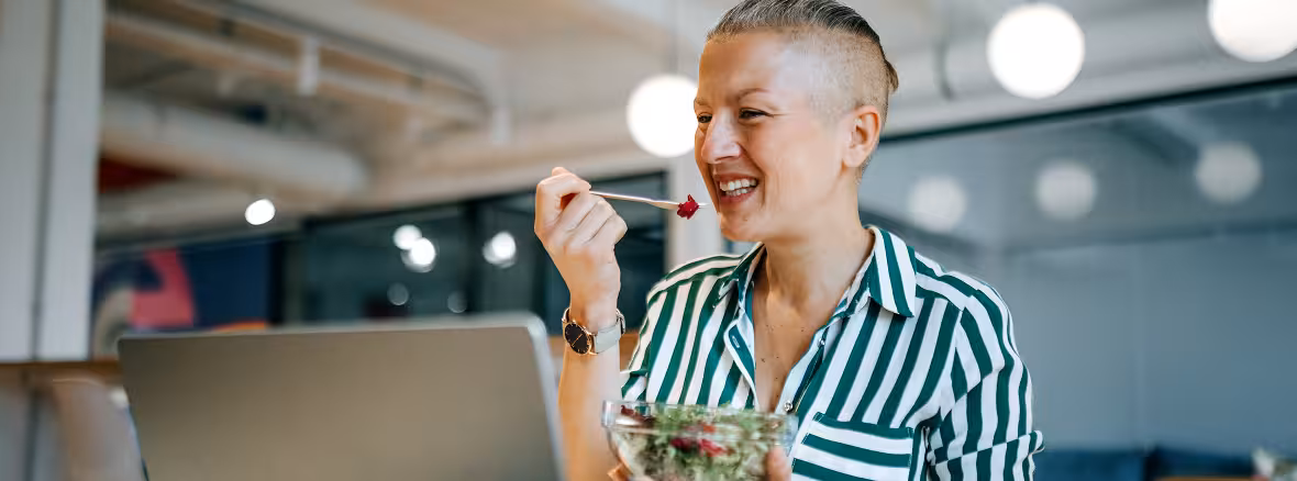 A person with short, grey hair and a striped shirt smiles while eating a salad from a clear bowl, with a laptop in front of them in an office setting.
