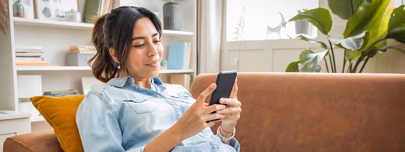 Woman sitting on a brown couch looking at her smartphone with a slight smile.